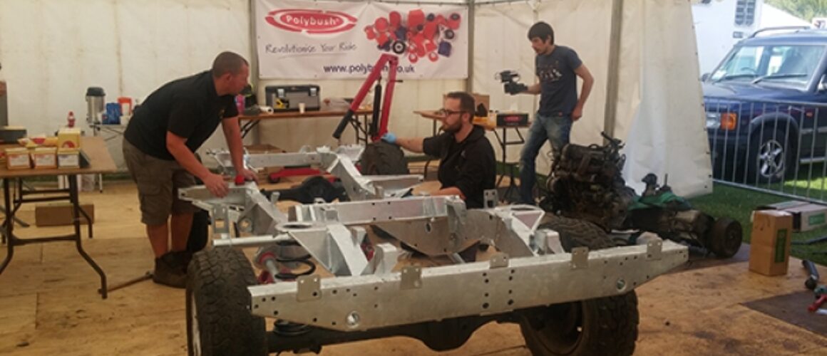 Three men work on a car chassis in a workshop marquee with various car parts and a camera setup; a banner with "polybush" hangs in the background.