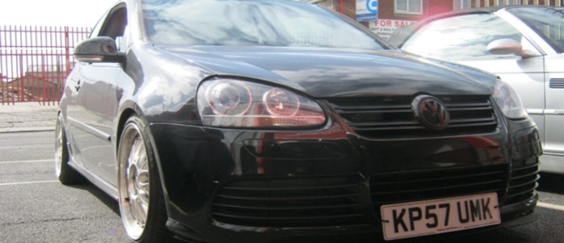 A low-angle view of a black Volkswagen car, parked on a street. The car features a distinctive front grille and round headlights, with a visible number plate reading "kp57 umk".
