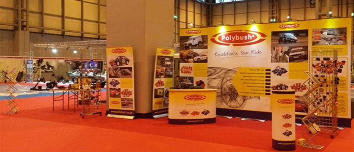 Exhibition stand showcasing polybush automotive products, featuring banner stands with images and information, surrounded by various car parts and enhancements in a well-lit hall.