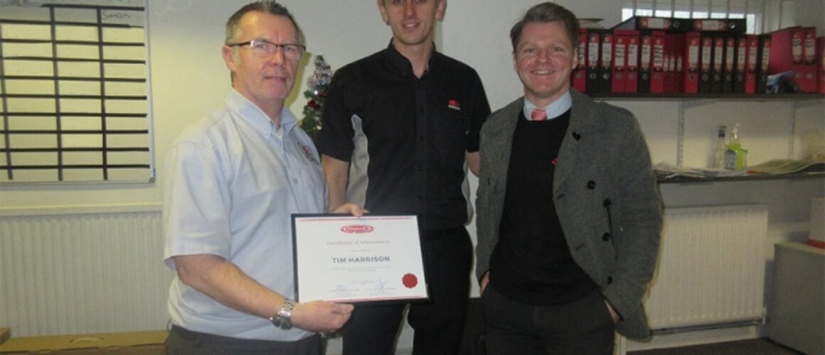 Three blokes in an office environment, grinning as the taller chap in the middle brandishes a certificate. In the backdrop, there are shelves filled with files.