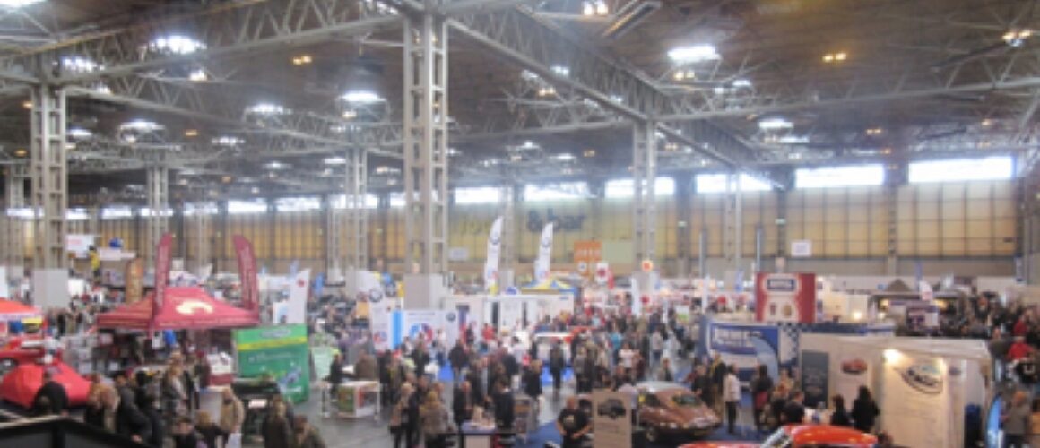 A busy indoor exhibition with various stalls, displays, and a throng of visitors. There are cars on show, promotional banners, and different trade stands in a large hall with high ceilings.
