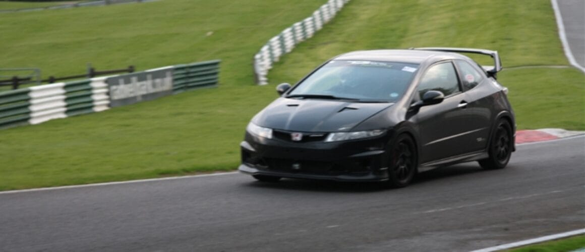 A black sports car racing on a curved circuit with lush green turf on the sides and white fencing in the background.