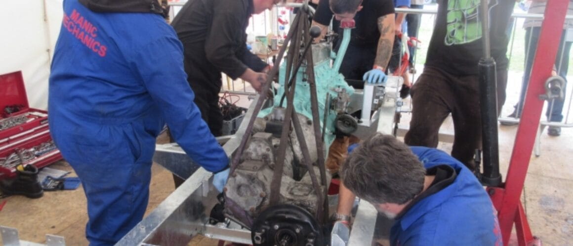 Five mechanics collaborate to fit an engine into a vehicle chassis in a workshop marquee, encapsulated by tools and parts.