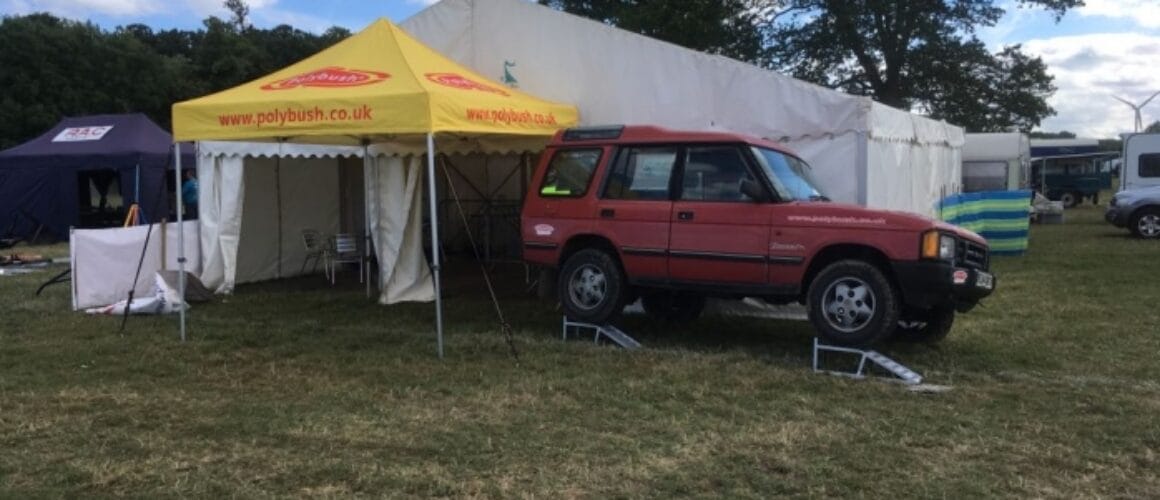 A red land rover parked beside a commercial marquee with banners at an outdoor event, with other marquees and vehicles in the background under a cloudy sky.