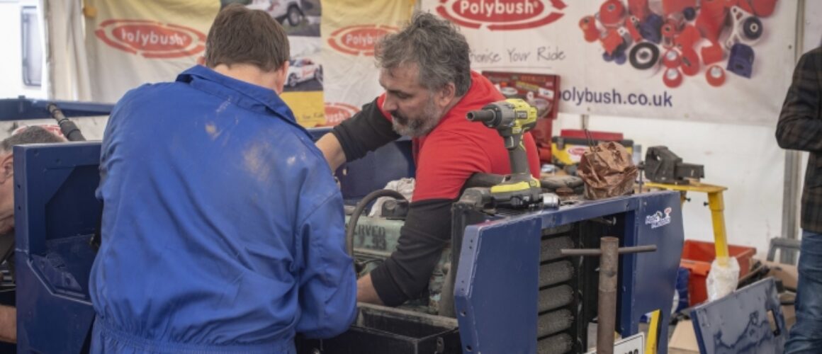 Two mechanics in blue coveralls working on a car engine in a marquee at an outdoor event, with a banner for "polybush" in the background.