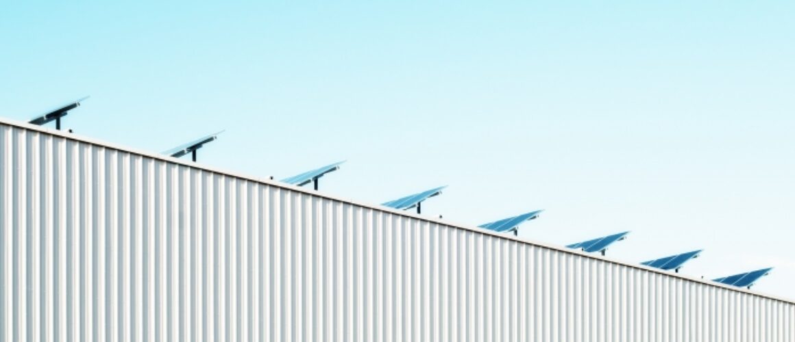 A minimalist image of a white metal building roofline against a clear blue sky, adorned with evenly spaced small wind turbines.