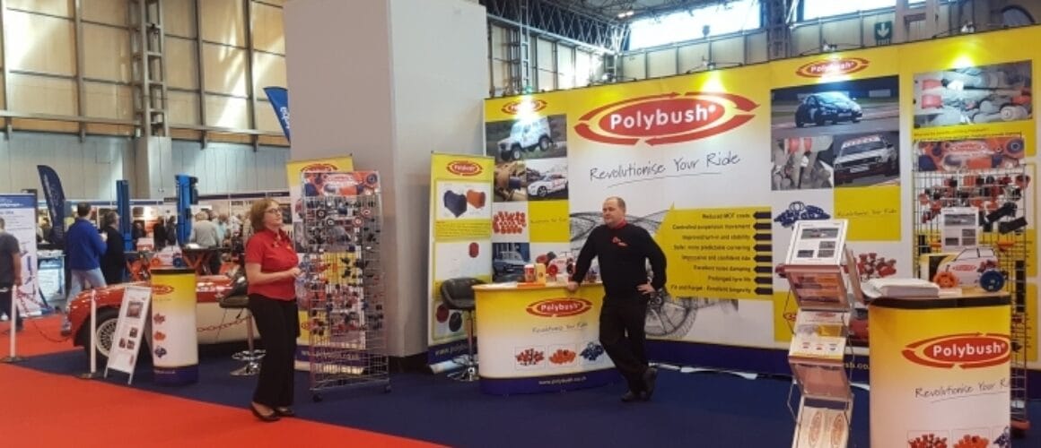 A trade show stand for "polybush" featuring automotive parts with two representatives, one male and one female, standing next to promotional displays in a brightly lit venue.