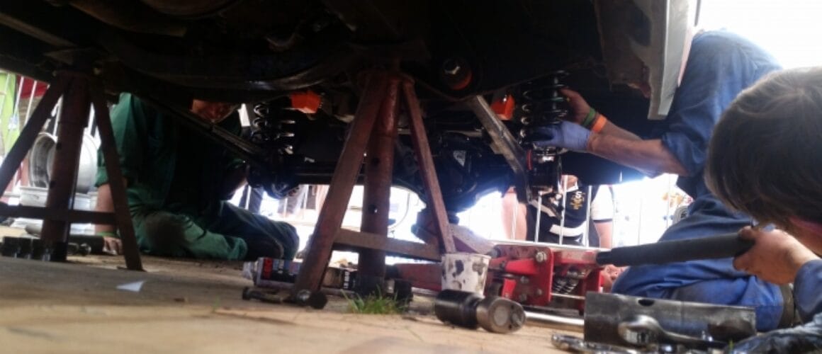 Three mechanics work beneath a hoisted car, utilising tools to mend or inspect its underside. The emphasis is on their hands and apparatus, with particulars of the vehicle's suspension visible.