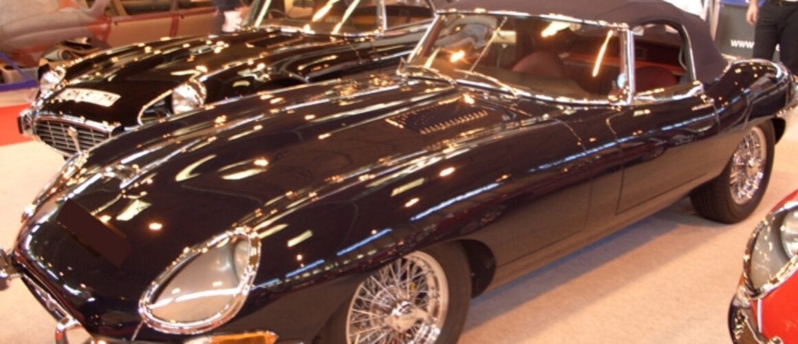 Two shiny classic Jaguar E-type cars displayed at a car exhibition, featuring sleek black and red models with chrome detailing and wire wheels.