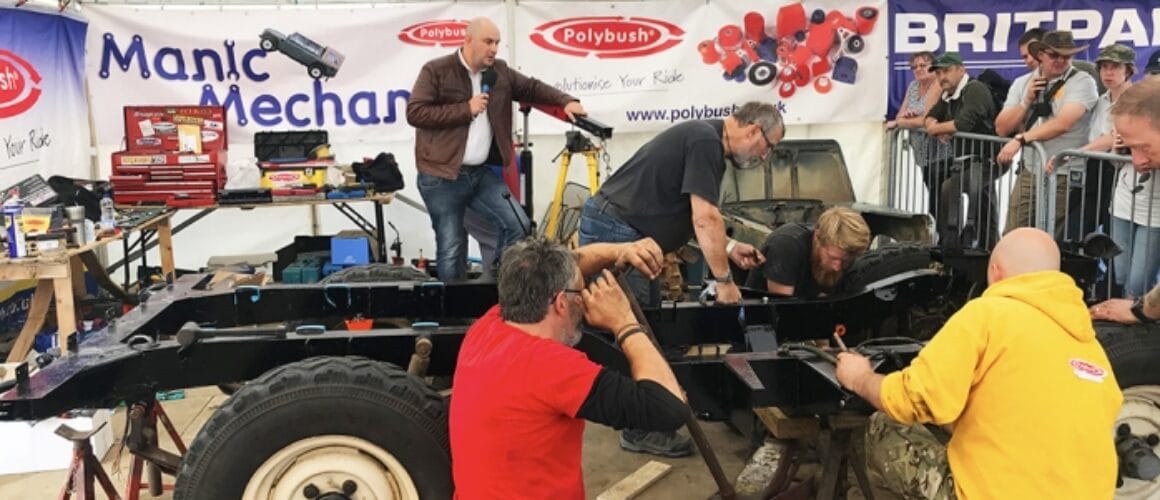 A group of individuals are observing a mechanic work on a vehicle chassis at an outdoor event beneath a marquee with various car parts and sponsor banners displayed in the backdrop.
