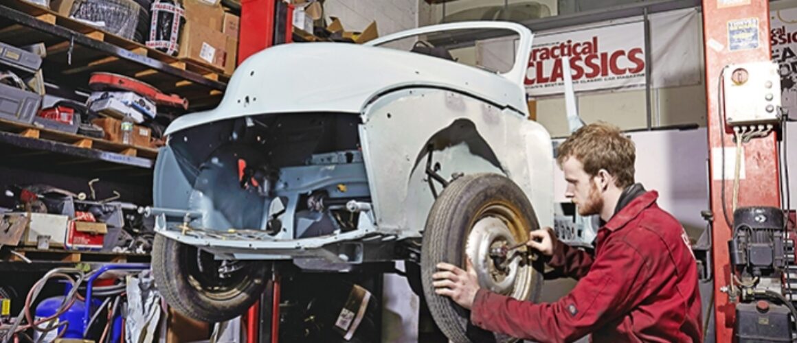 A mechanic in a red boiler suit works on restoring a classic white car in a cluttered workshop filled with shelves of parts and tools.