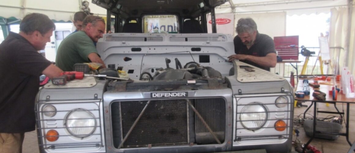 Three men working on a dismantled Land Rover Defender in a marquee workshop, surrounded by tools and vehicle parts.