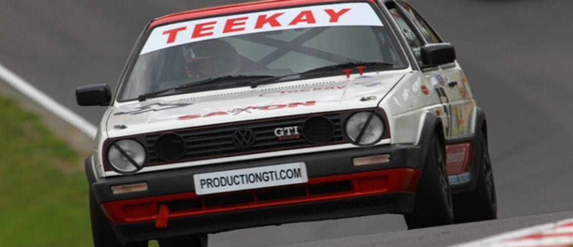 A vintage Volkswagen Golf GTI race car speeds on a racecourse, prominently featuring sponsor logos "teekay" and "productiongti.com" on a grey day.