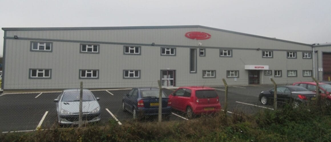 A large industrial grey building with a red logo on top, a car park in front with various cars, and some greenery in the foreground on a foggy day.