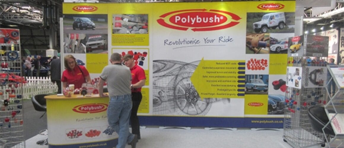 Two people stand at the polybush exhibition booth with car part images and text, at an automotive trade fair. A banner with the slogan "revolutionise your ride" is displayed prominently.