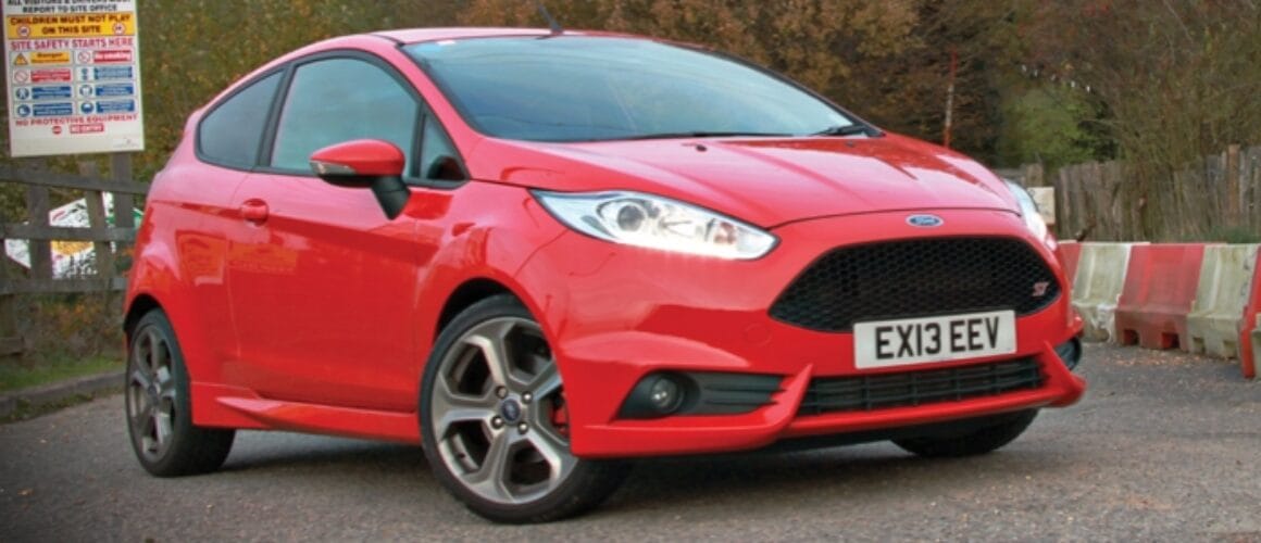 A bright red Ford Fiesta parked outside, boasting a sleek design with stylish alloy wheels, situated in front of a fence with parking notices.