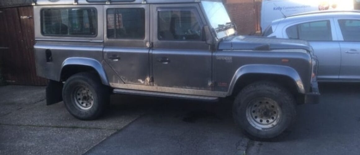 A grey land rover defender parked diagonally on a narrow street, with a wooden garage and another car in the background.