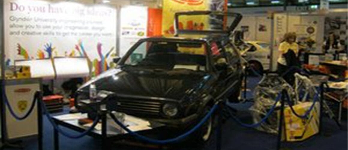 A black car on display at a crowded trade fair stand with various promotional materials and signs, including one that asks, "do you have a big idea?