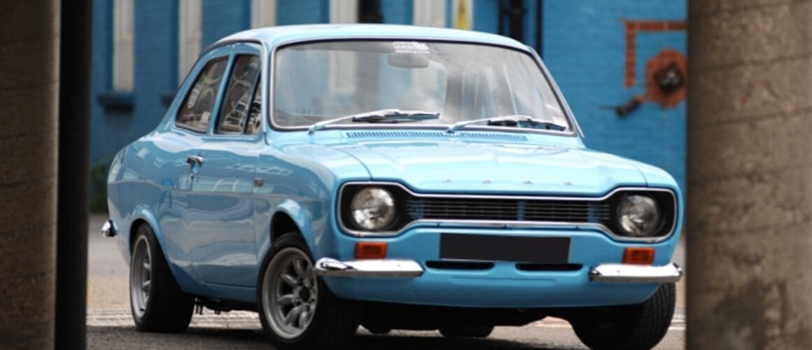 A classic blue Ford Escort car parked on a street, observed between two pillars, in an urban setting with brick walls and a hint of graffiti.