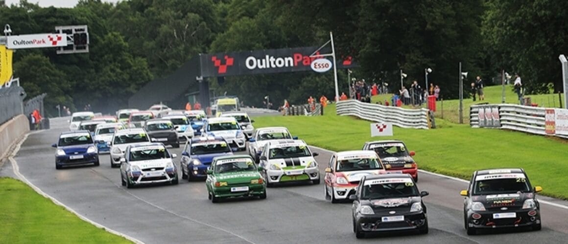 A motor racing event with a variety of cars in multiple rows at the start of a damp circuit, spectators line the barriers, and flags and branding visible in the background.