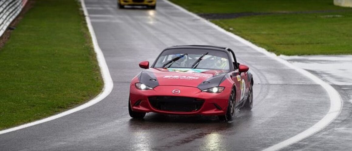 A red Mazda convertible leads a race on a damp circuit, followed by several other sports cars, including a yellow one, at a motorsport event.