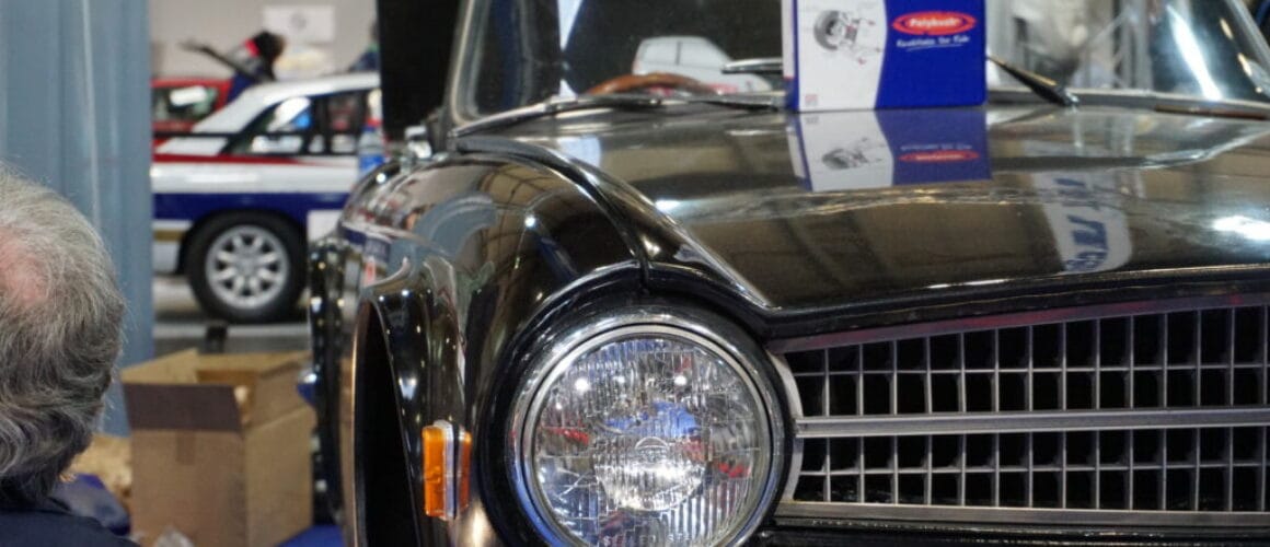 Close-up of a vintage black car with a reflective chrome grille and round headlights at an indoor exhibition, surrounded by event attendees and exhibits.