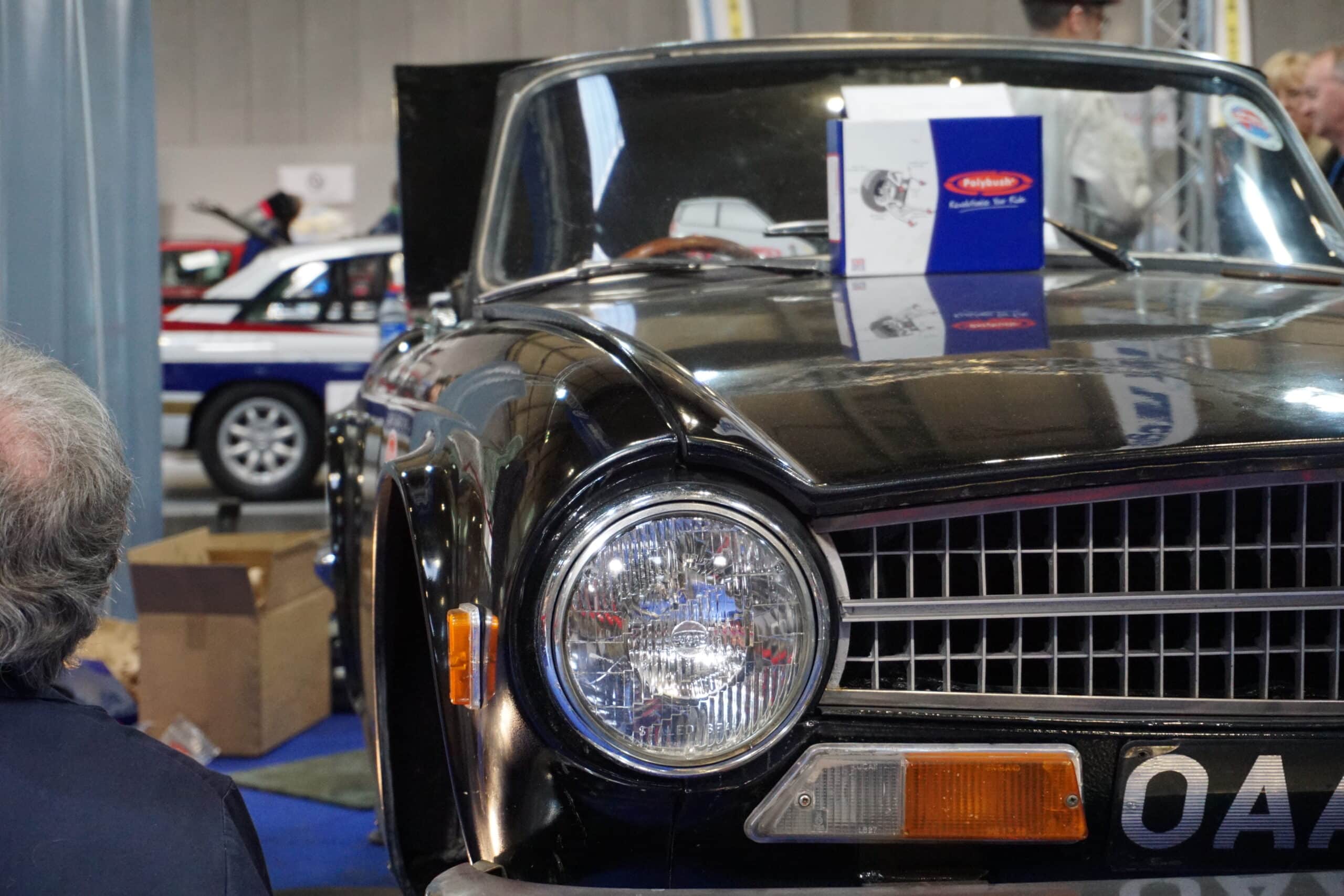 Close-up of a vintage black car with a reflective chrome grille and round headlights at an indoor exhibition, surrounded by event attendees and exhibits.