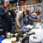 Two individuals in a workshop arrangement at an exhibition, with one working on a dismantled car axle and the other observing and taking photos. Various tools and car parts are visible around them.