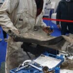 A mechanic in a grubby overall holds a large, dusty car part in a workshop with tools scattered on a table and a classic car in the backdrop.