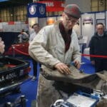 A bloke in a cap and dungarees works on a car part in a bustling exhibition hall, whilst another bloke holding a microphone observes him.