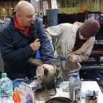 Two blokes work on a car part at an exhibition stand, one observing while the other uses tools, with automotive products and a classic car in the background.