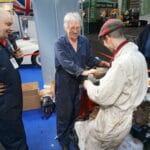 Three blokes interact at a mechanical workshop surrounded by car components. One in a blue uniform laughs, another in spectacles exchanges an object with a third man in a dirty white overall.