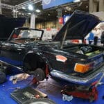 A classic black convertible car with its bonnet open and parts laid out on the ground, being repaired at an indoor exhibition area with people in the background.