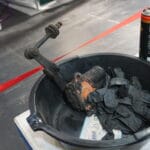 A black bucket containing a used angle grinder, a rusty crank handle, and mucky work gloves, placed on a grey floor with a red line, alongside a tin of lubricant spray.