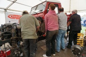 A group of individuals observing a mechanic work on a red tractor engine at an outdoor event under a marquee with a "polybush" banner in the backdrop.