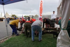 A group of individuals collaborating to put together or mend wooden structures outdoors at a festival, with tents and promotional banners nearby.