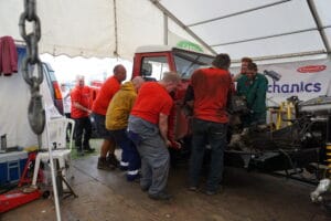 A group of mechanics in red shirts working on a dismantled red car inside a marquee workshop, crowded around the open engine bay.