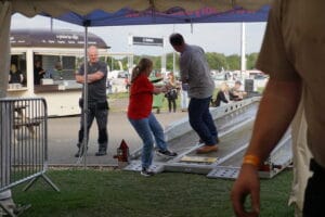 A woman in red and a man race slot cars on a track at an outdoor event, with spectators watching nearby.