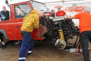 Four men work on a red off-road vehicle in a marquee area, concentrating on its exposed engine and suspension. One observes, whilst three are actively engaged in the maintenance task.