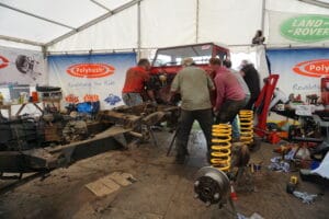 A group of individuals labour on dismantling a red Land Rover in a marquee filled with car parts and tools at an outdoor event.