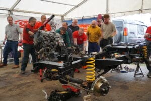 A group of individuals collaborating to fit an engine into a vehicle chassis in a marquee workshop area, with various tools and equipment visible.