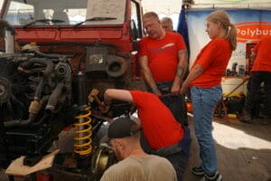 Three individuals in orange shirts labour on large vehicle parts in a marquee at an open-air market, with one person crouched closely inspecting the engine.