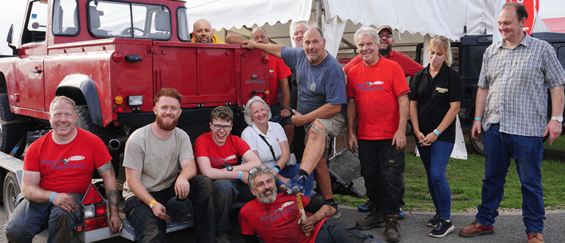 A group of ten cheerful individuals, some in red shirts, pose around a classic red lorry in a field with tents in the background, creating a merry, communal atmosphere.