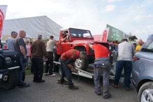 People gather around a red off-road vehicle being unloaded from a trailer at an outdoor event, with marquees and a blue sky in the background.