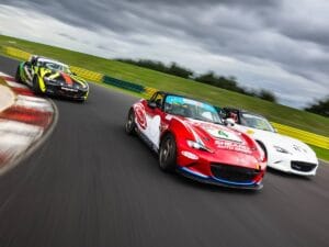 Three Mazda sports cars racing on a circuit, with the nearest car in red featuring prominent sponsorship logos, against a backdrop of clouds.