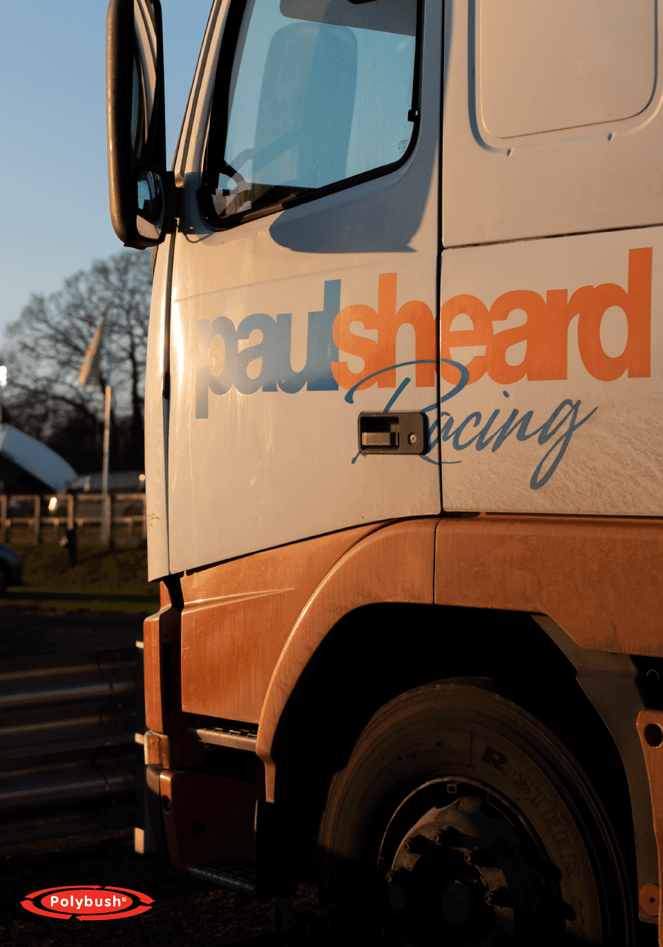 Side view of a dusty orange lorry with "paul sheard racing" printed on its door, lit up by early morning sunlight, with a blurred background of railings and flags.