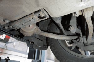 Undercarriage view of a car showing the suspension system and part of the exhaust, focusing on a rusty track control arm and other components.