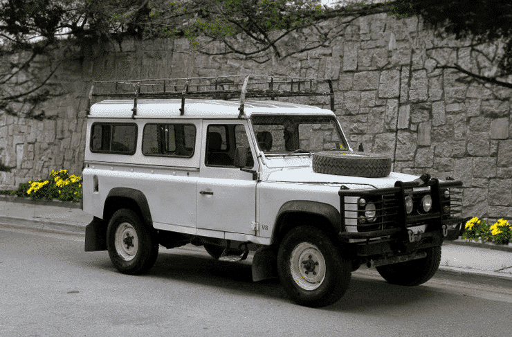 A white, rugged Land Rover Defender with a roof rack and spare tyre mounted on the front, parked beside a stone wall on a street lined with blooming yellow flowers.