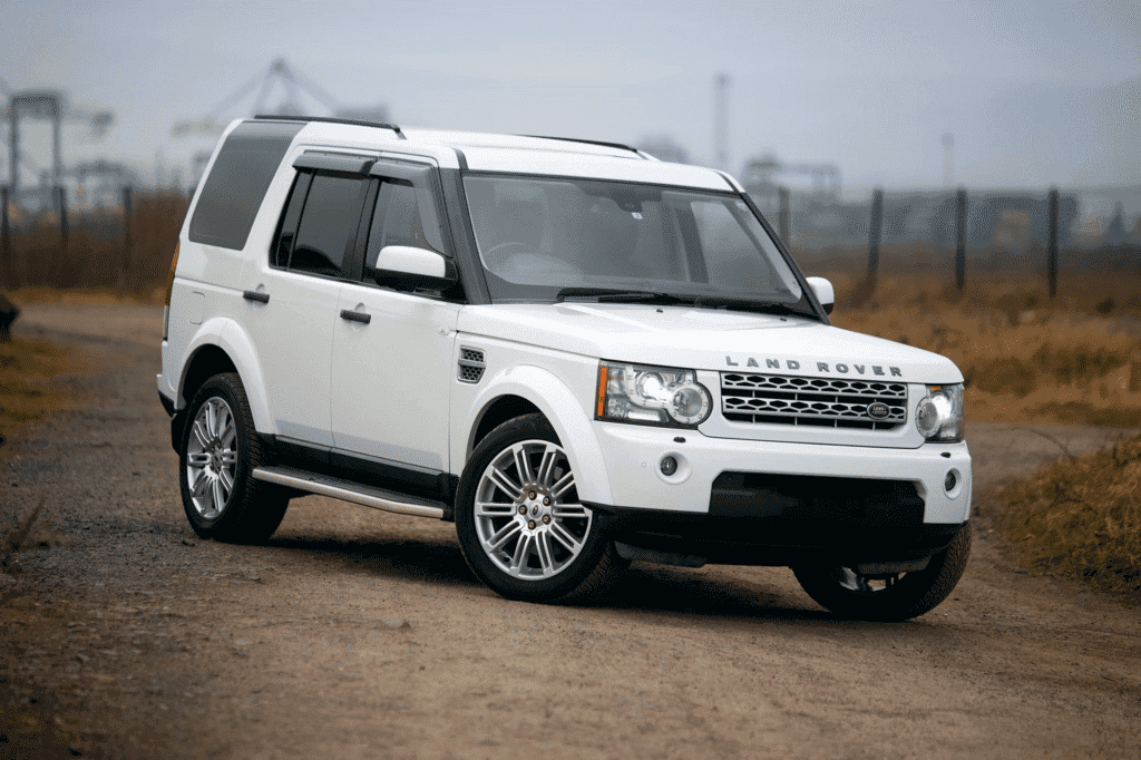 A white Land Rover Discovery parked on a gravel road with a grassy field and foggy background. The vehicle is prominently displayed with a clean and glossy exterior.