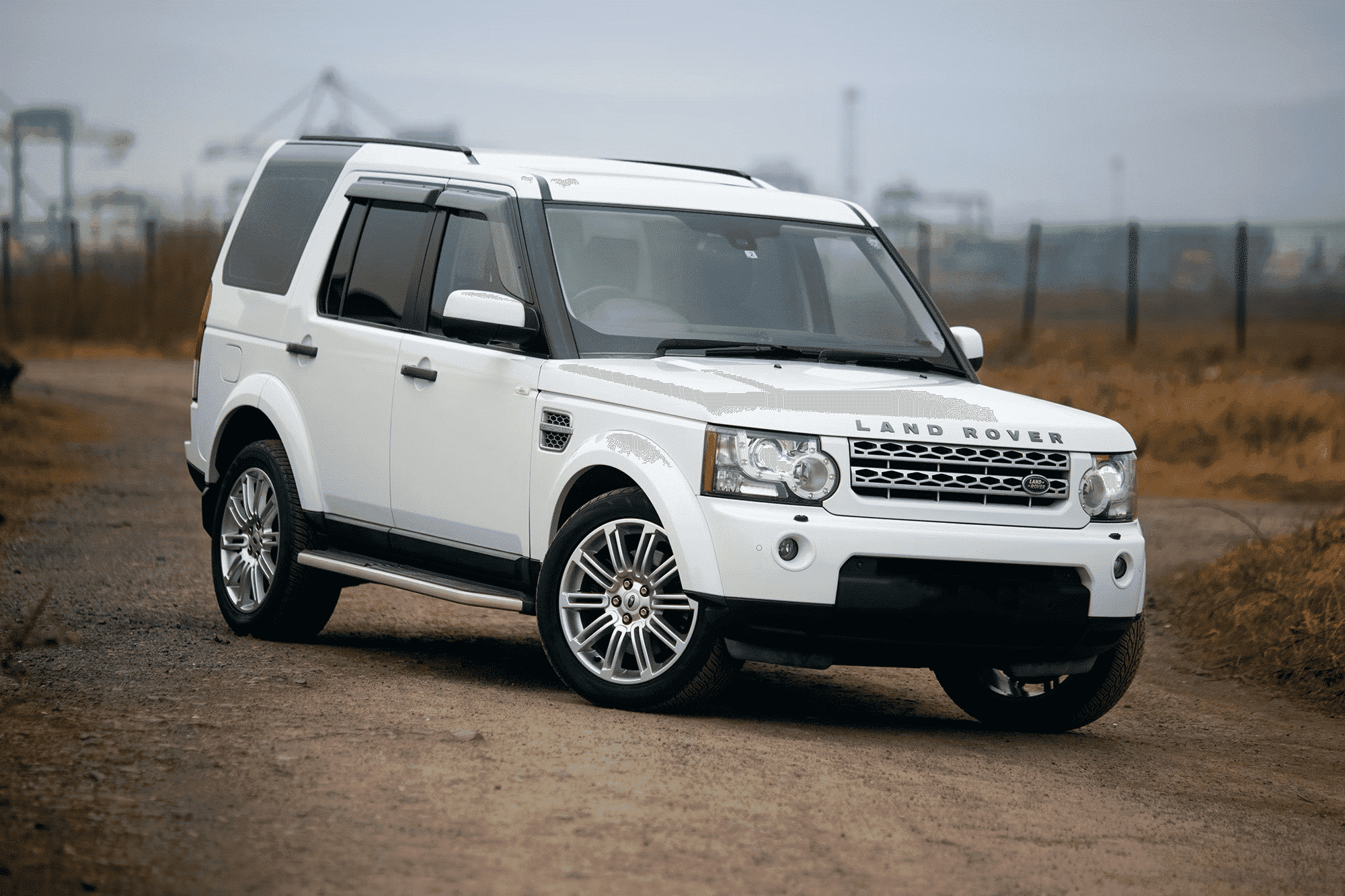 A white Land Rover Discovery parked on a gravel road with a grassy field and foggy background. The vehicle is prominently displayed with a clean and glossy exterior.
